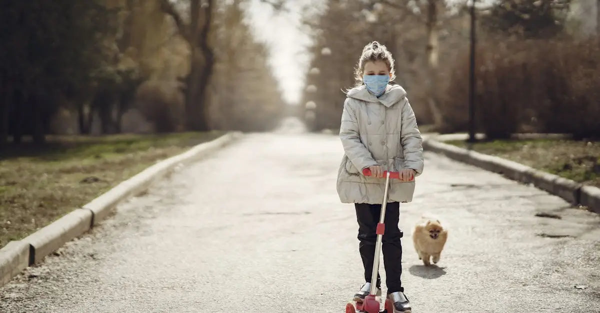 Girl wearing mask rides scooter with small dog on park path outdoors in autumn.