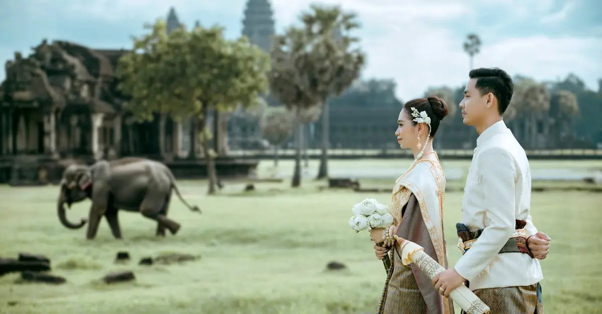 Bride and groom in traditional attire with bouquet near elephant and ancient temple in grassy field